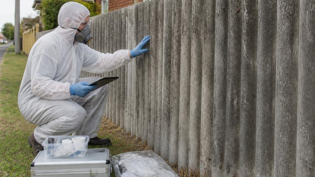 A licensed asbestos removalist in full white protective coveralls, respirator and gloves carefully inspecting a weathered fibro cement sheet fence on a residential property, with a clipboard and testing kit visible nearby